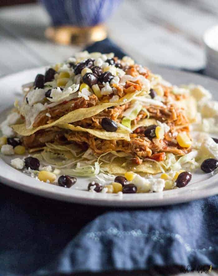 An image of the Slow Cooker Salsa Chicken Tostadas recipes on a white plate.  There are 3 tostadas stacked on top of each other with black beans, corn and lettuce on the plate.  There is a navy blue napkin underneath the plate.
