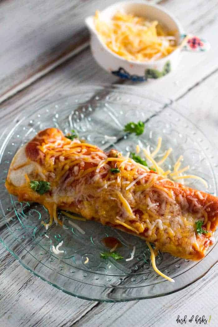 An image of the Instant Pot chicken enchiladas recipe on a clear glass plate. There is a small bowl with shredded cheese in the background of the image.